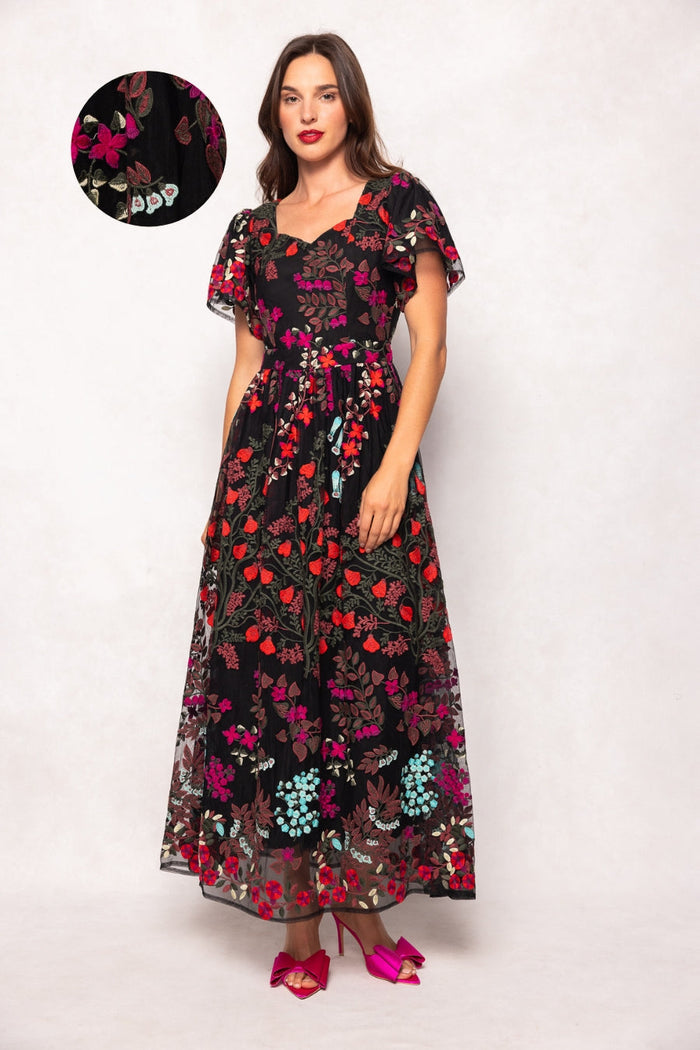 A woman wears the Flora Dress in Moonlit Blossom by Ivy City Co—a black, floor-length gown adorned with vibrant, intricately embroidered florals. The dress features short flutter sleeves and a belted waist. She stands against a light background, and there is a close-up detail showcasing the elaborate embroidery on the fabric.