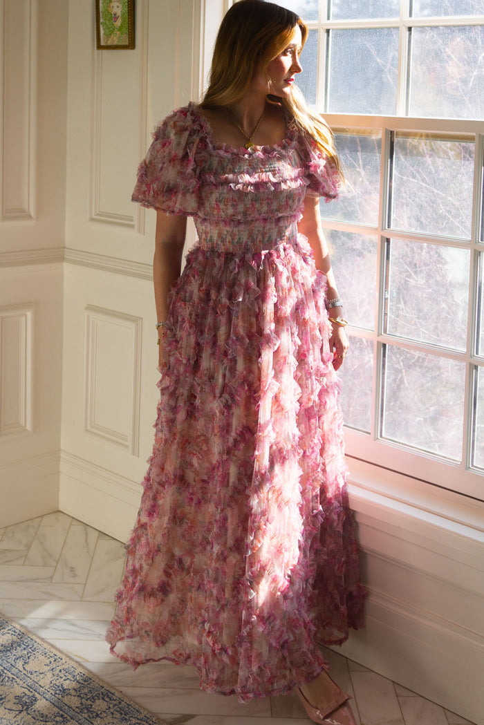 A woman stands by a sunlit window in a white paneled room, wearing the Ivy City Co Florentina Dress in Alluring Floral—ruffled layers and dress pockets highlighting its timeless femininity as she gazes outside.