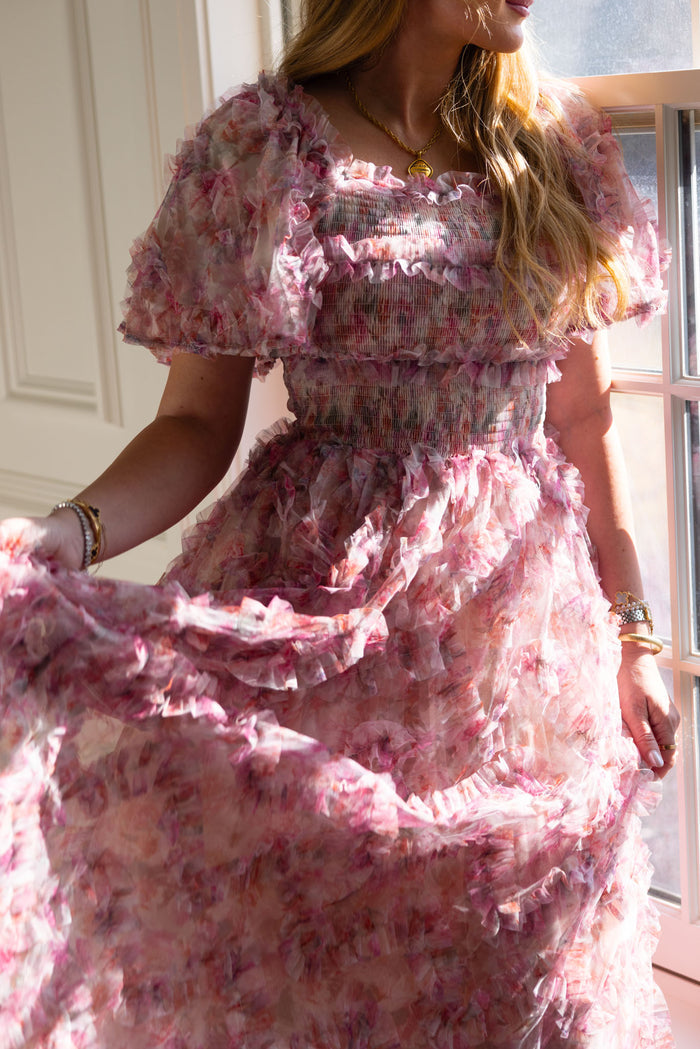 A woman in sunlight wears Ivy City Co’s Florentina Dress in Alluring Floral—its pink ruffled print and puff sleeves evoke timeless femininity. She shows off its flowy skirt, her wavy hair loose and gold jewelry catching the light.
