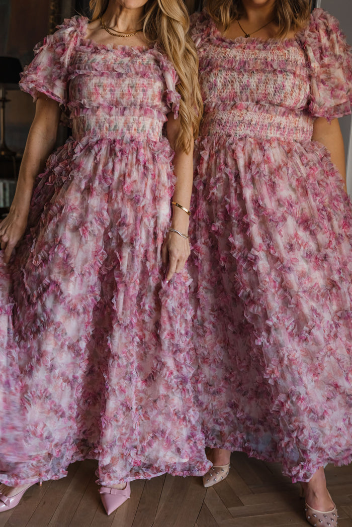 Two women stand side by side in Ivy City Co’s Florentina Dress in Alluring Floral—pink, ruffled maxi styles with smocked bodices and puff sleeves. Both wear bracelets and pointed-toe heels, radiating timeless femininity in a softly lit indoor setting.