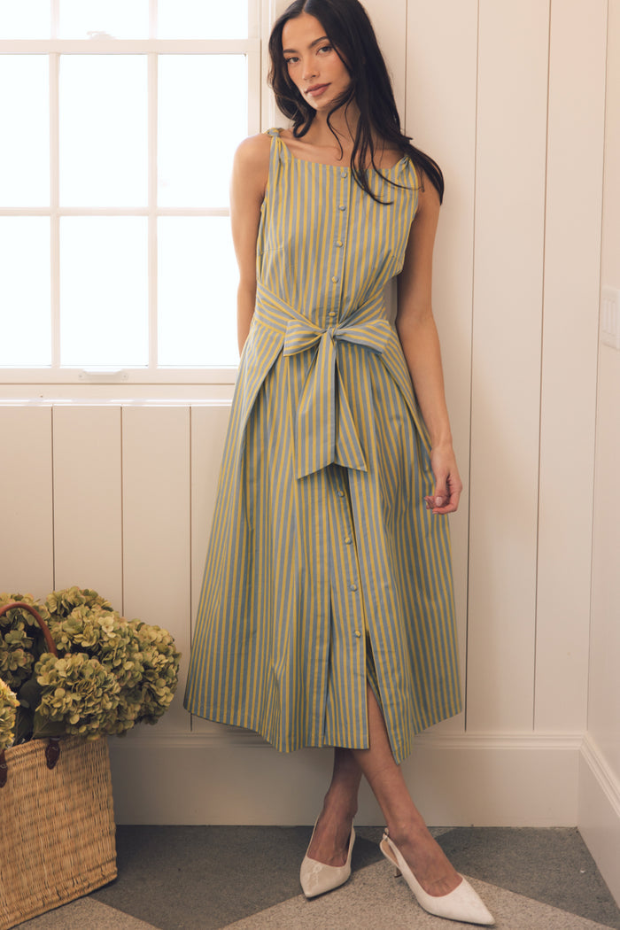 A woman stands indoors by a window, wearing the Ivy City Co Glen Dress in Green Stripe with a bow at the waist and white heels. A basket with green hydrangeas sits on the floor beside her.
