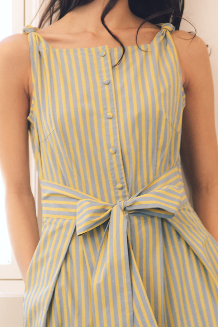 A woman wearing the Glen Dress in Green Stripe by Ivy City Co, a sleeveless nursing-friendly dress with vertical green and white stripes, front buttons, and a tied waist bow, shown from shoulders to knee.