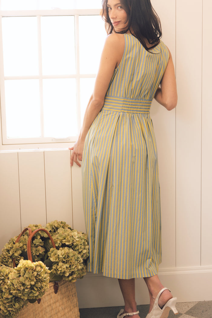 A woman stands indoors by a window, looking over her shoulder, wearing the Ivy City Co Glen Dress in Green Stripe. She is near a large basket filled with green hydrangeas and pairs the sleeveless, ankle-length dress with light heels.