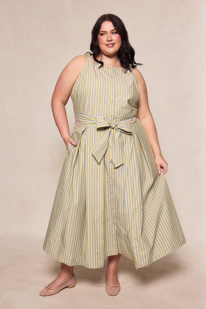 A woman wears the Ivy City Co Glen Dress in Green Stripe, a nursing-friendly midi with front buttons and a waist tie. She poses with one hand in her pocket and the other holding her skirt, paired with nude flats.