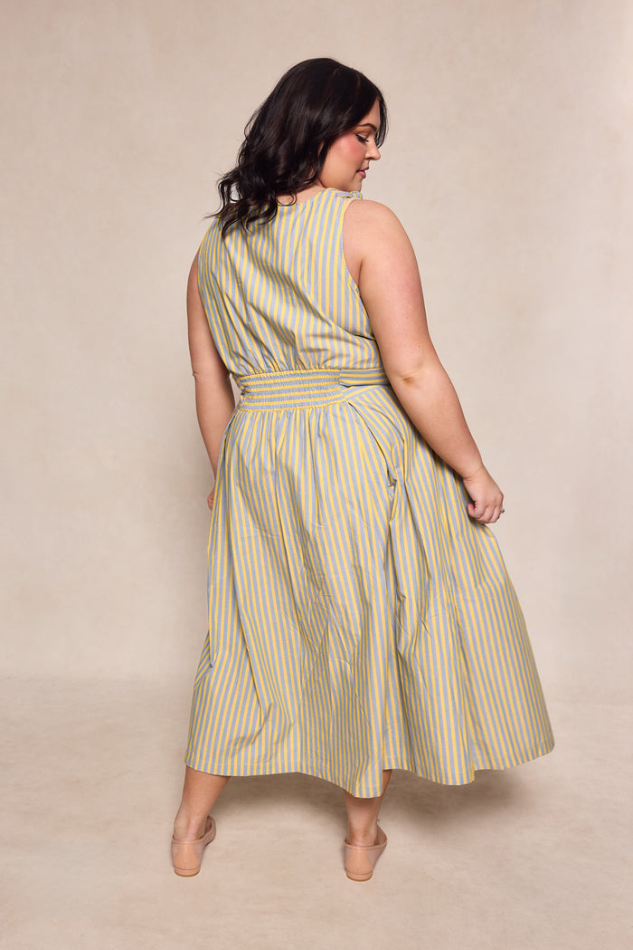 A woman with wavy dark hair models the Ivy City Co Glen Dress in Green Stripe, a sleeveless, cotton ankle-length dress. She stands with her back to the camera, looking over her shoulder against a neutral background.