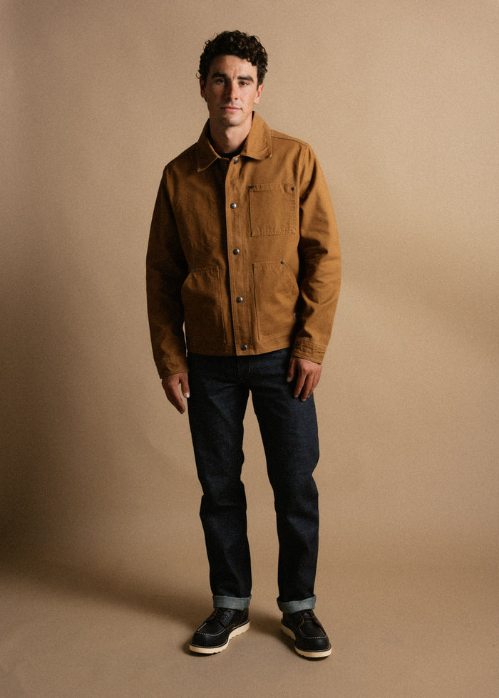 Man in brown jacket and dark jeans stands against a beige background, looking at the camera with a neutral expression.