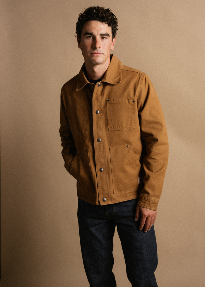 Man in brown jacket and jeans stands against a beige background, hands in pockets, looking confidently at the camera.