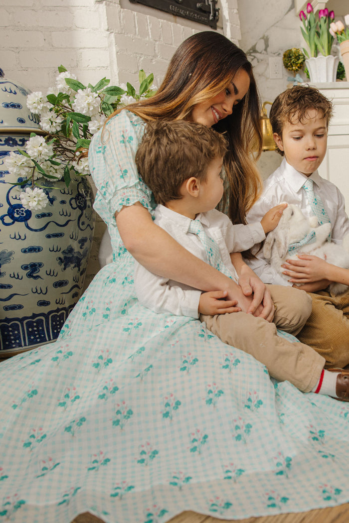 A woman wearing the Ivy City Co Jessie Dress in Bloom Gingham sits on the floor with two young boys, one holding a white rabbit. Nearby are a large blue and white vase, green plants, and a white brick fireplace with tulips.