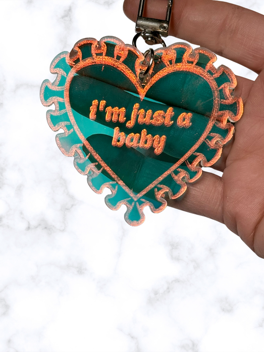 Iridescent heart acrylic keychain with "Just a Baby" design by Adorrible, featuring a shiny, colorful surface and a metal keyring attachment.