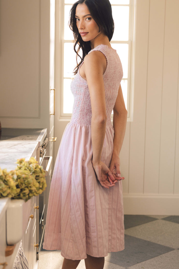 A woman with long dark hair stands in a softly lit room, wearing the Ivy City Co Karla Dress in Pink Striped Gingham—a maternity friendly style with a fitted bodice and pleated skirt—offering effortless elegance.