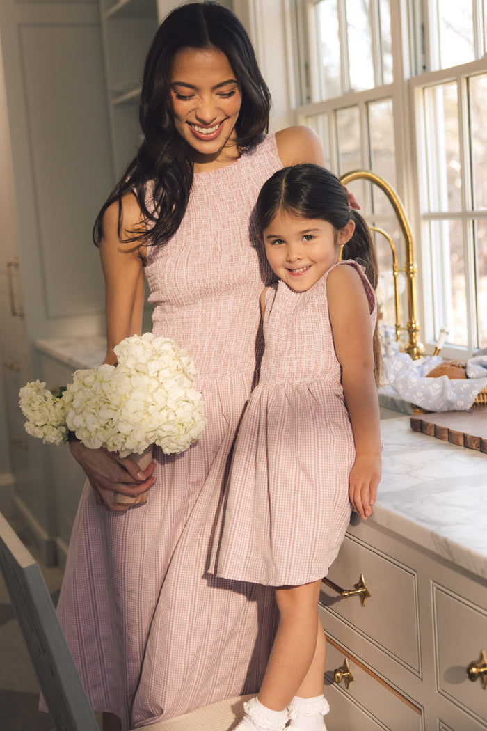 A smiling woman in the Ivy City Co Karla Dress in Pink Striped Gingham holds white flowers, standing with a young girl in matching dresses on a kitchen counter, both bathed in sunlight from a nearby window.
