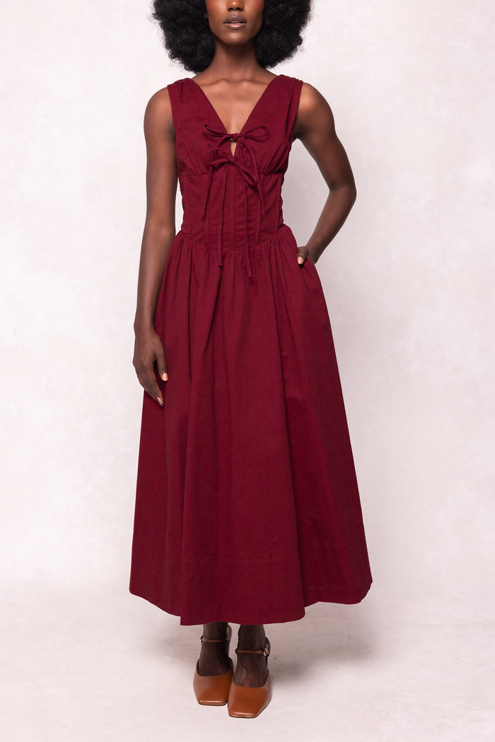 A woman models the Ivy City Co Kayla Dress in Burgundy—a sleeveless, midi-length dress with a fitted bodice, corset waist, front bow, and flared skirt—posing against a light background with one hand in her pocket and brown shoes.