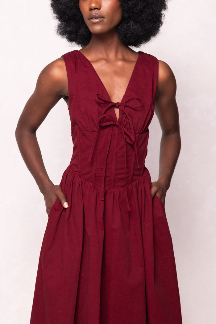 A woman wears the Kayla Dress in Burgundy by Ivy City Co—a sleeveless midi dress with a deep v-neck, bow tie detail at the chest, and pockets—standing against a light background.