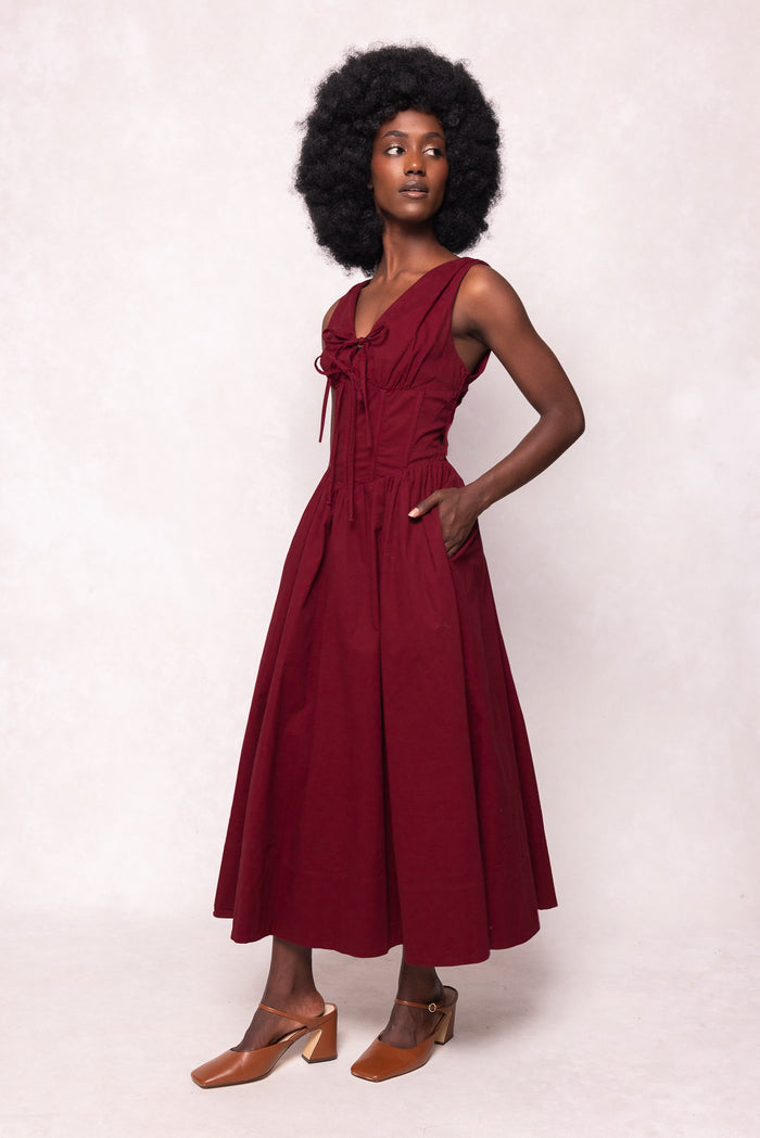 A woman with a natural afro wears the Ivy City Co Kayla Dress in Burgundy—a sleeveless midi dress with a corset-style waist and pockets. She poses against a light background, one hand in her pocket, paired with brown heeled shoes.