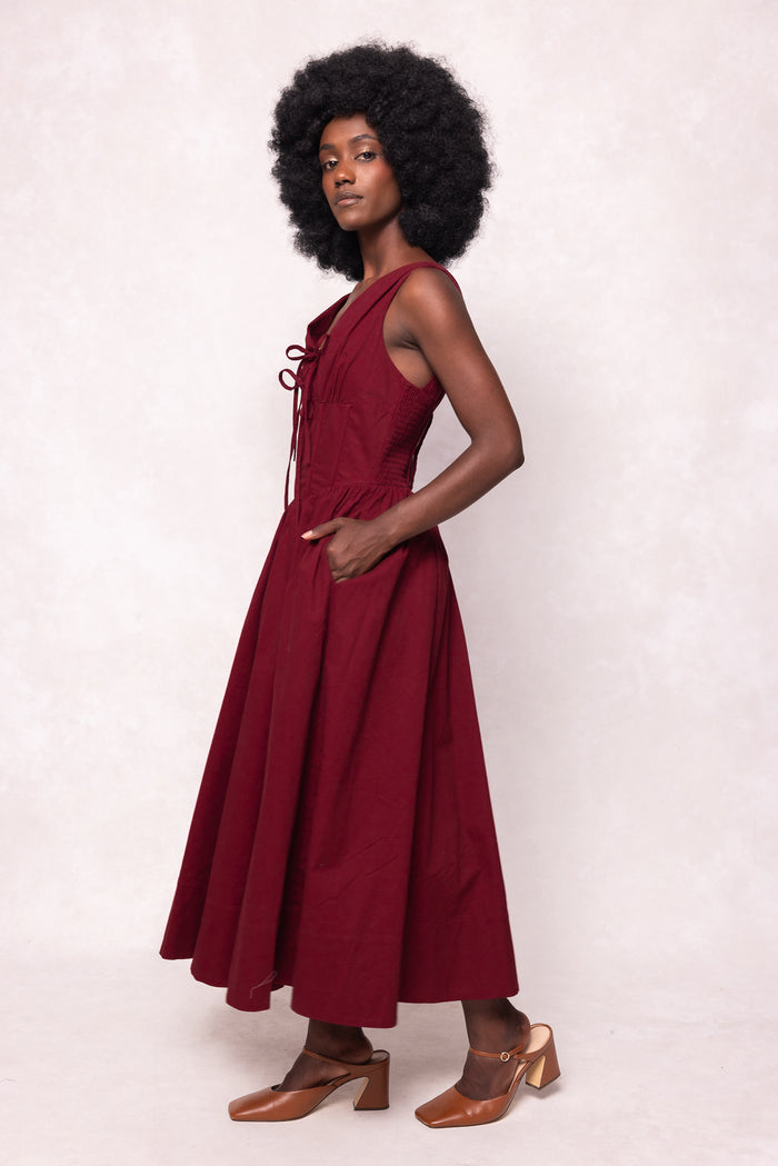 A woman with an afro hairstyle models the Ivy City Co Kayla Dress in Burgundy—a sleeveless midi dress with a corset waist and v-neckline. She stands sideways, one hand in her pocket, wearing tan heels against a light background.