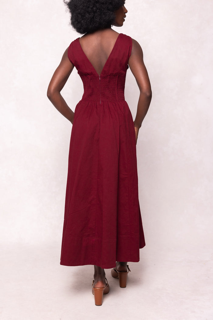 A woman faces away, modeling the Ivy City Co Kayla Dress in Burgundy. This sleeveless midi features a v-neckline, corset-style ruched bodice, and flowing skirt. She pairs it with brown heeled sandals against a plain light background.