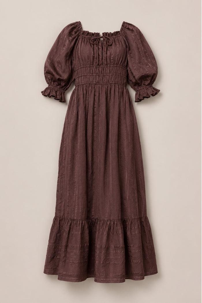 The Ivy City Co Maleah Dress in Warm Cocoa features a long maxi silhouette with puffed sleeves, ruffled cuffs, a smocked bodice, tie detail at the neckline, and a ruffled hem. Laid flat on a neutral background, this nursing-friendly style offers both comfort and elegance.