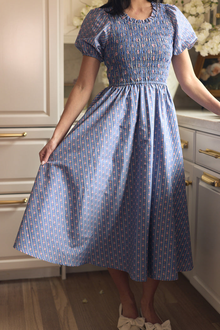 A woman stands in a kitchen wearing Ivy City Co’s Mariah Dress in Linear Print, a blue, short-sleeved, smocked, maternity-friendly dress with red and white patterns. She holds one side of the full skirt; light cabinets and flowers are behind her.