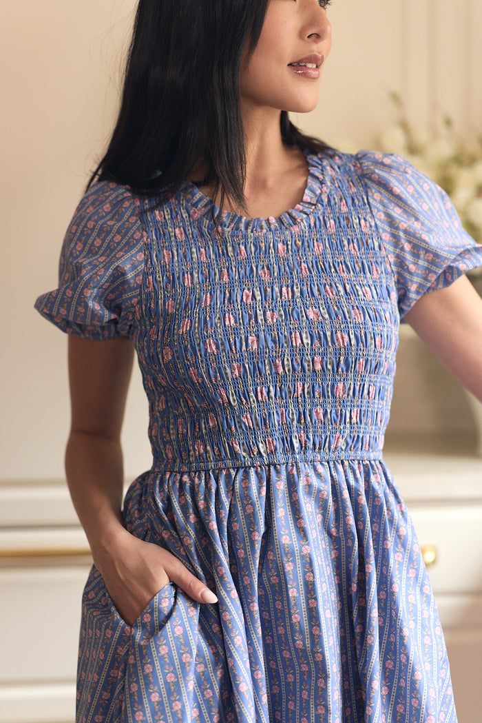 A woman in the Ivy City Co Mariah Dress in Linear Print, featuring short puff sleeves and a smocked bodice, stands partially turned with one hand in her pocket. Her long dark hair is down. The softly blurred background has light décor. Maternity friendly.