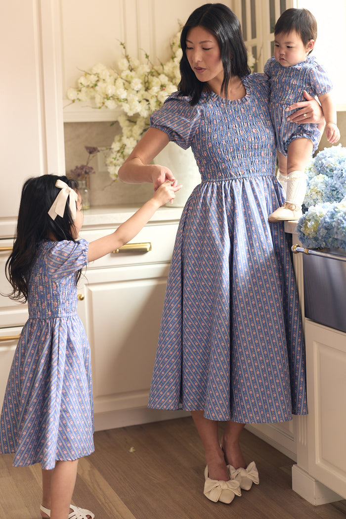 A woman in the Ivy City Co Mariah Dress in Linear Print stands in a kitchen with two children, all wearing matching blue patterned outfits. She holds a baby and gently touches hands with an older girl in a white bow.