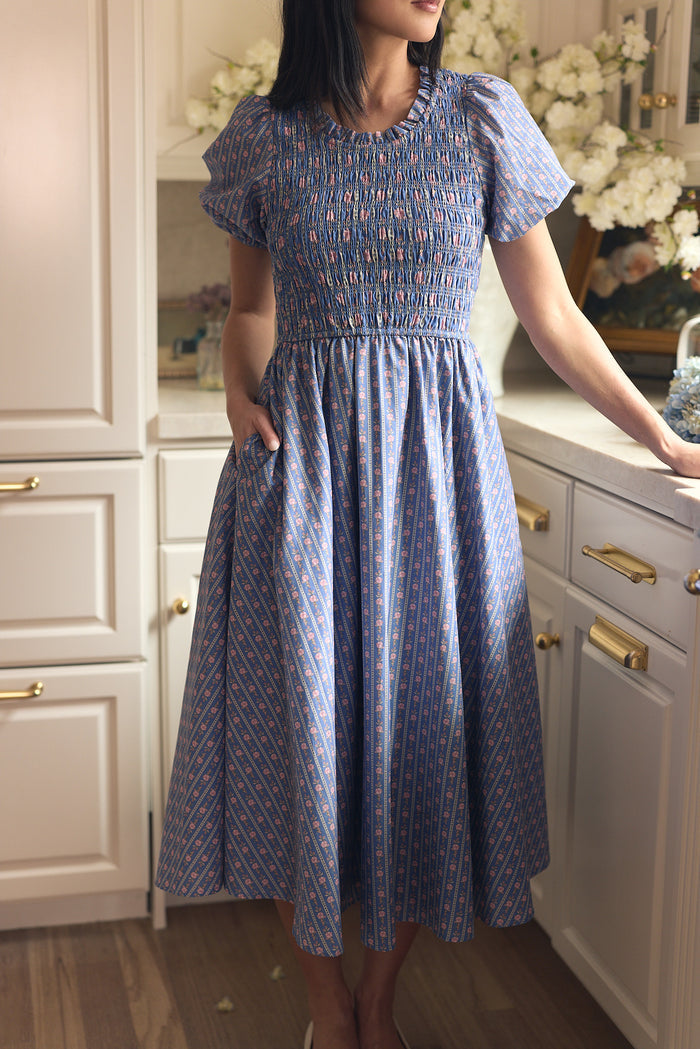 A woman in the Ivy City Co Mariah Dress in Linear Print, a maternity-friendly cotton dress with short puff sleeves, stands in a kitchen with white cabinets and brass handles, surrounded by white flowers. Her face is not visible.