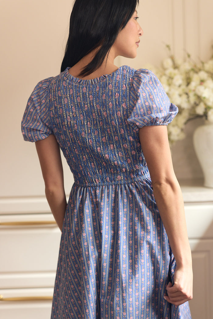 A woman with long dark hair, seen from behind, wears the Ivy City Co Mariah Dress in Linear Print—a blue, maternity-friendly style with puff sleeves and a smocked back—standing indoors by white flowers on a console table.