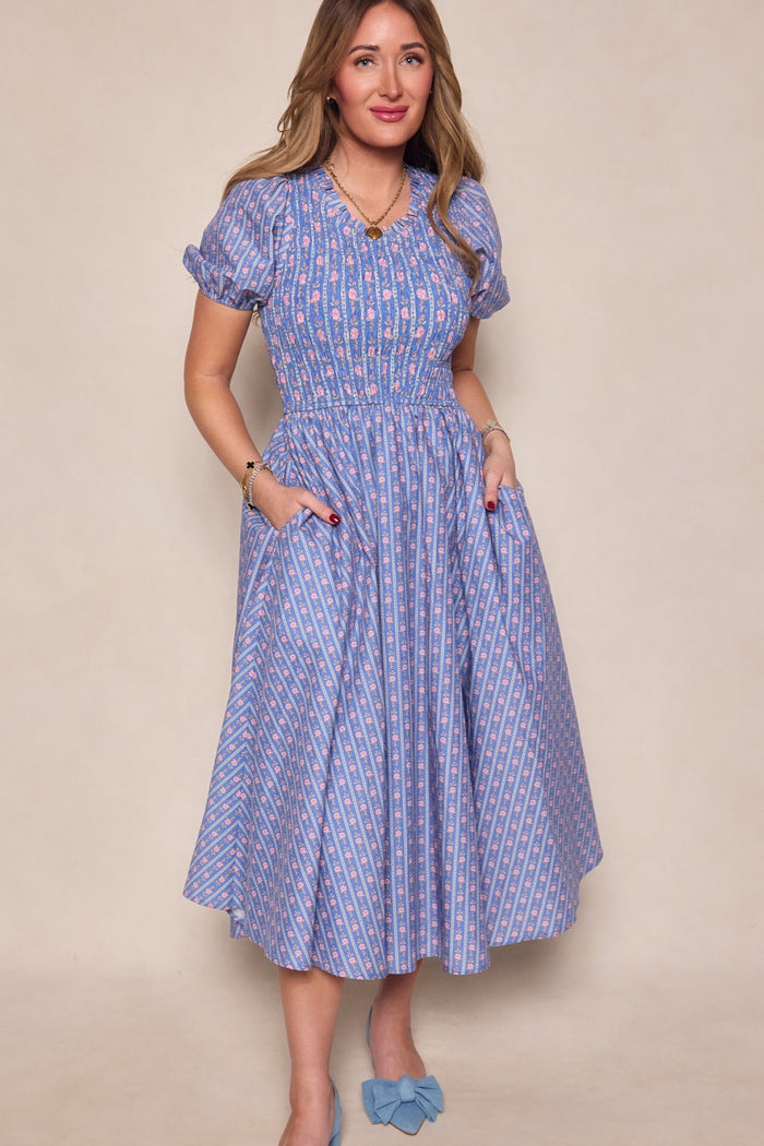 A woman with long brown hair smiles against a plain background wearing the Ivy City Co Mariah Dress in Linear Print—a blue patterned, short-sleeved, midi floral cotton dress with pockets. She pairs it with blue bow shoes and gold jewelry. Maternity friendly.