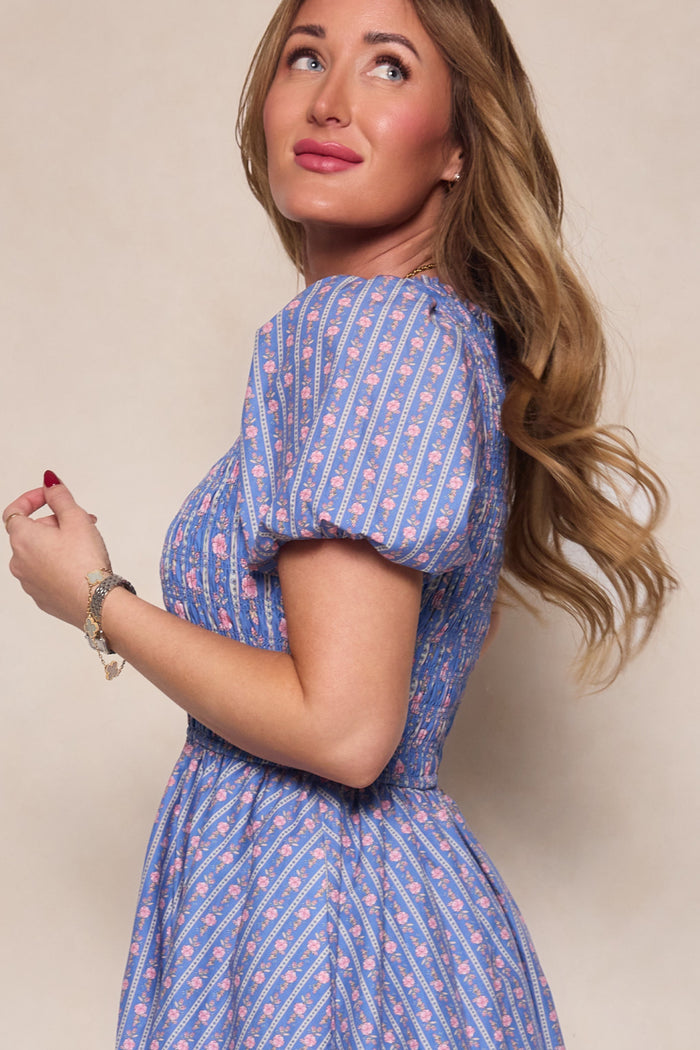 A woman with long wavy hair, wearing Ivy City Co's Mariah Dress in Linear Print, stands sideways against a neutral background and smiles softly, highlighting the dress's maternity-friendly style.
