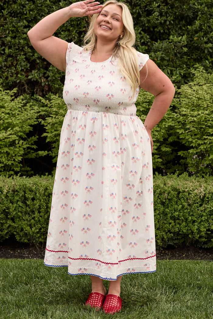 A smiling woman with long blonde hair stands on grass, saluting with her right hand while wearing the Ivy City Co Marlow Cap Sleeve Dress in Star Spangled and red shoes. Green bushes are in the background.
