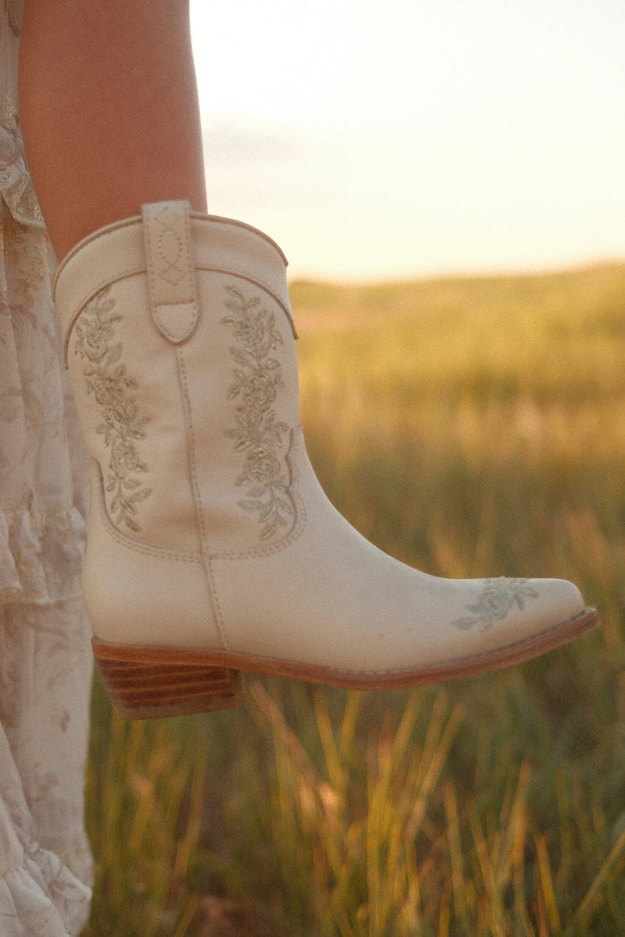 A close-up of the Mini Avery Boot in Cream by Ivy City Co, featuring floral embroidery, worn outdoors in a grassy field at sunset—a charming footwear choice for any adventure.