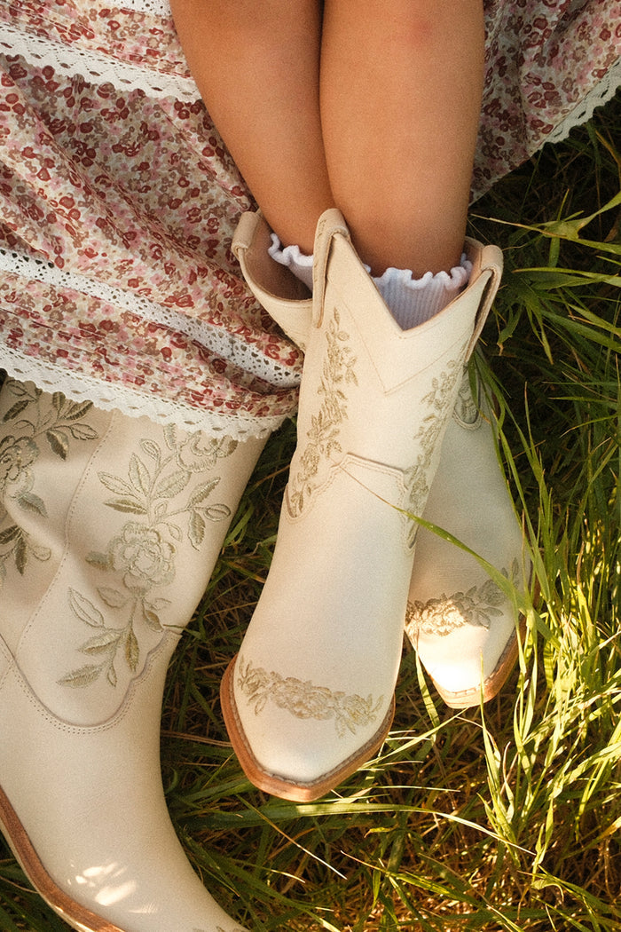 A person in a pink floral dress and Ivy City Co’s Mini Avery Boot in Cream sits on green grass, sunlight illuminating the lace details and embroidered flowers on the boots.