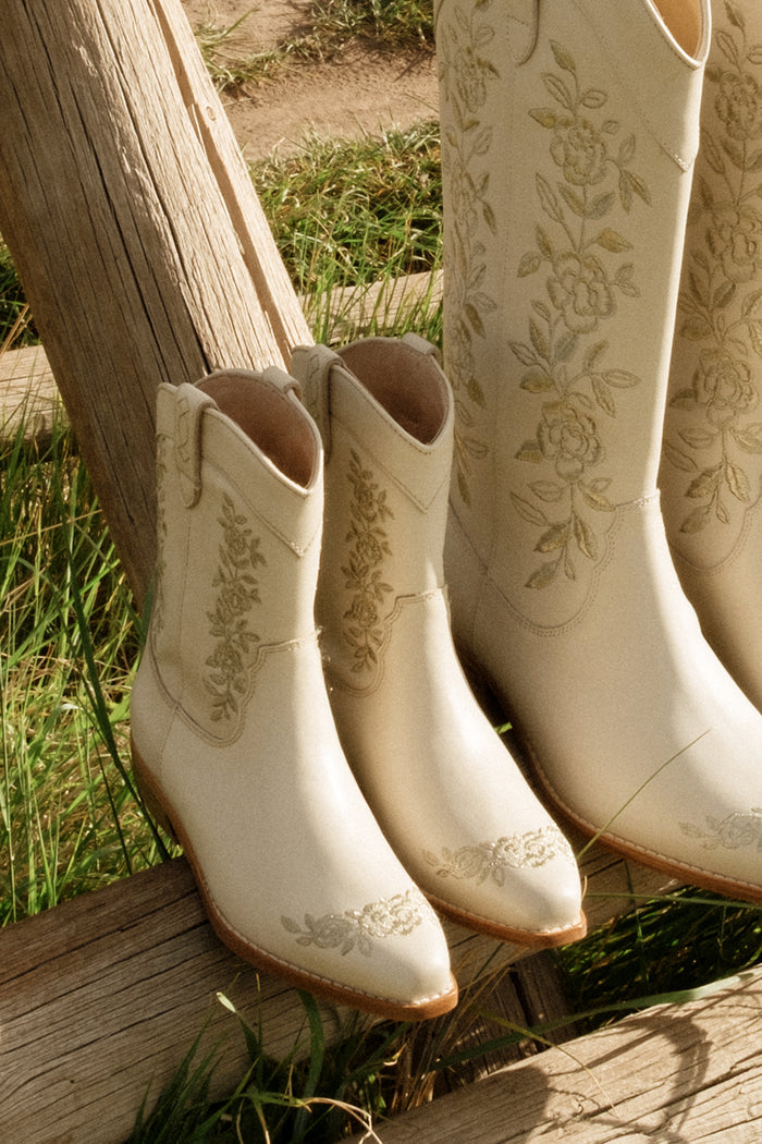The Ivy City Co Mini Avery Boot in Cream for toddlers, featuring floral embroidery, sits next to matching taller cream boots on a wooden bench outdoors with green grass in the background.