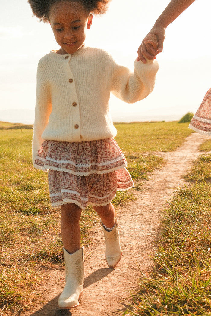 A young girl in a white sweater, floral skirt, and Ivy City Co’s Mini Avery Boot in Cream walks hand-in-hand with an adult along a sunlit path, surrounded by green grass and a bright, natural backdrop.