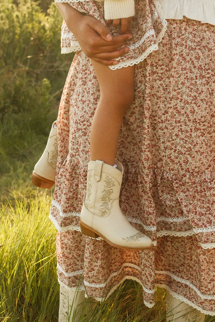 A child wears the Ivy City Co Mini Avery Boot in Cream and is held by an adult in a long, floral lace-trim skirt, standing on grass in warm sunlight.