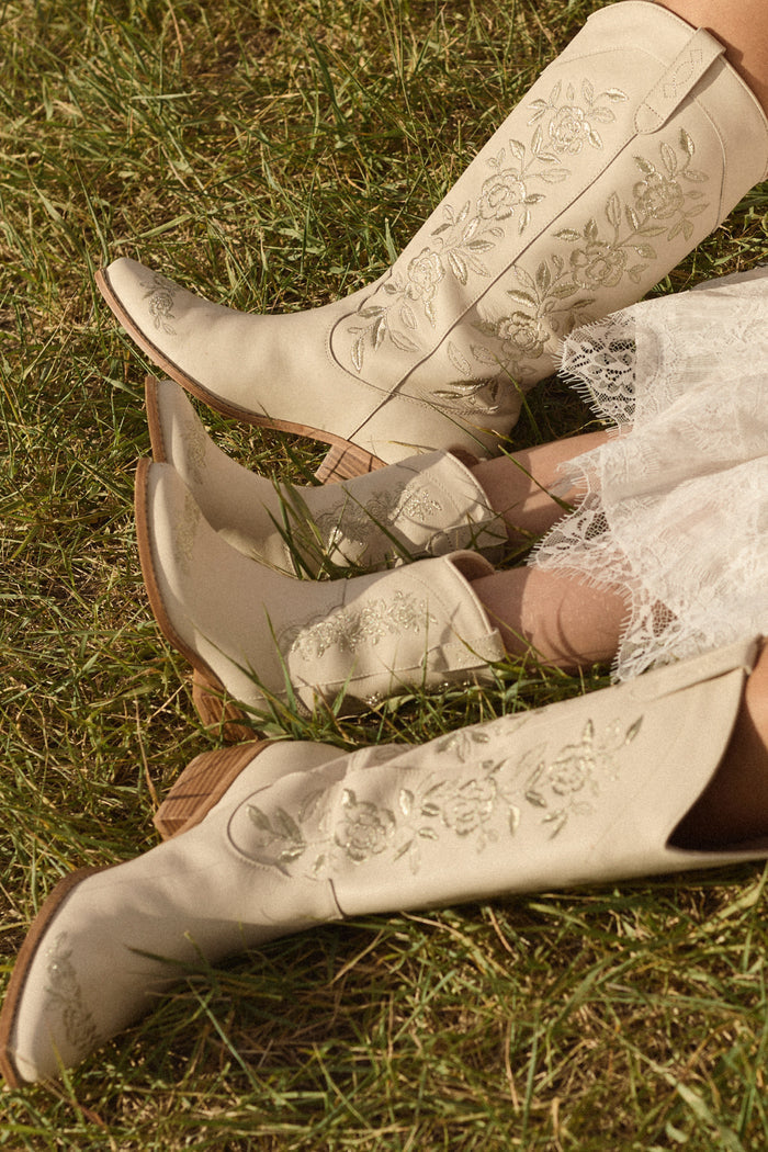Two people in matching cream boots, including the Ivy City Co Mini Avery Boot in Cream for toddlers, and white lace dresses sit on grass with legs extended and their boots side by side.