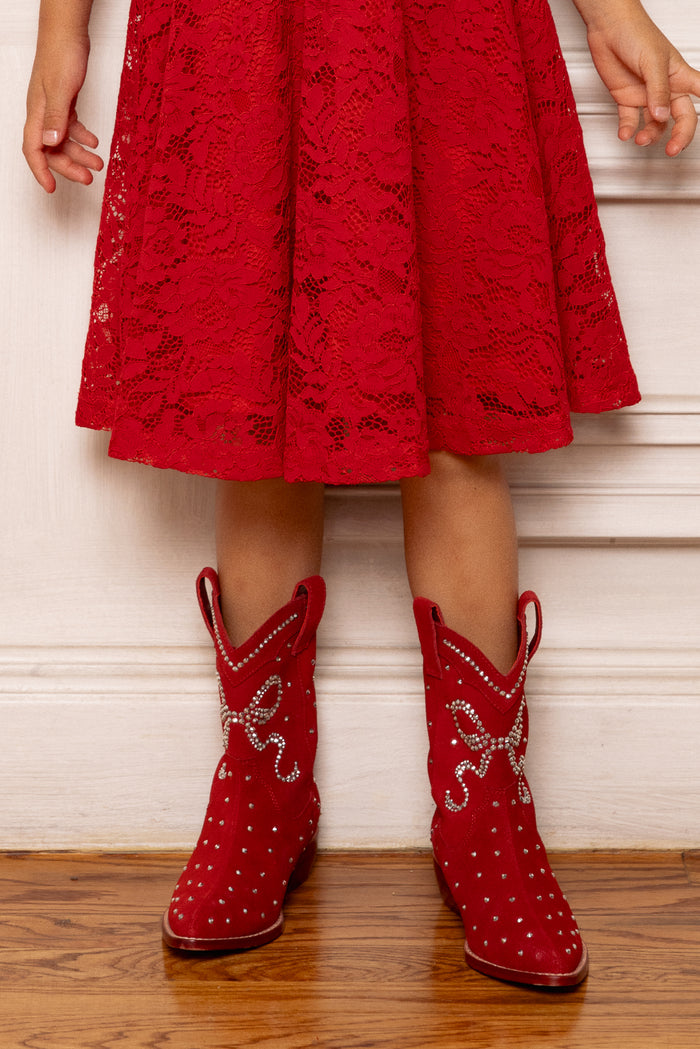 A child in a red lace dress and Ivy City Co's Mini Channing Bootie in Red stands on a wooden floor before a white wall, creating the perfect mommy-and-me twinning moment.