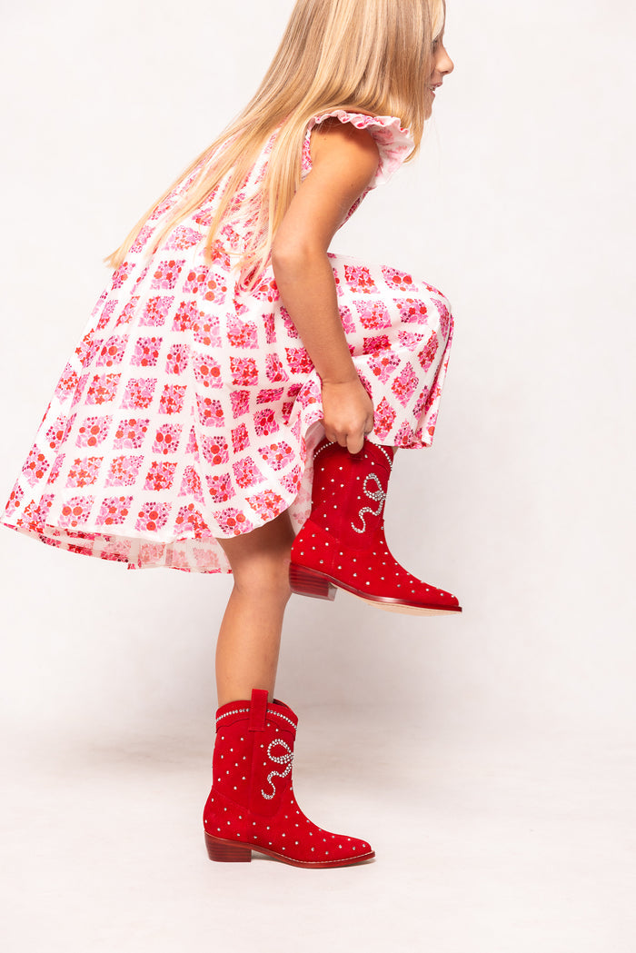A young girl in a pink and white floral dress bends to adjust her Ivy City Co Mini Channing Bootie in Red kids cowboy boots with decorative designs. Her long blonde hair falls over her shoulder against a plain white background.