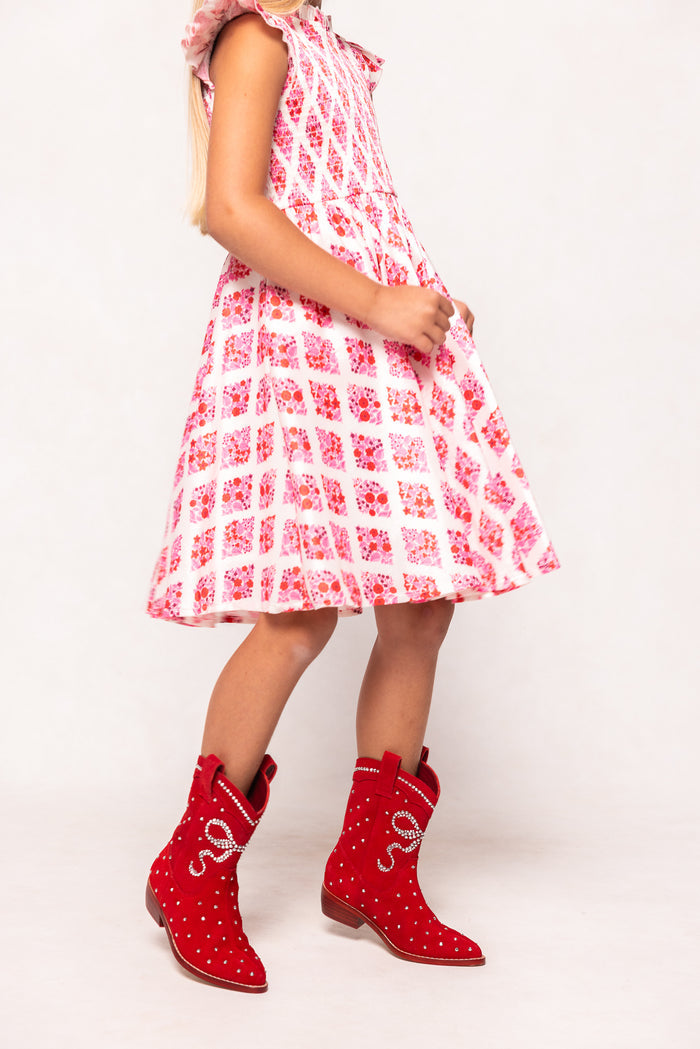A young girl wears a pink and white patterned dress with ruffled sleeves and Ivy City Co's Mini Channing Bootie in Red, standing against a plain light background—a perfect look for mommy-and-me twinning.