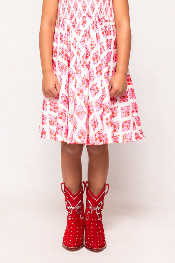 A young child wears a pink and white dress with Ivy City Co's Mini Channing Bootie in Red, featuring white detailing, against a plain white background—perfect for adorable mommy-and-me twinning moments. The child's face is not visible.