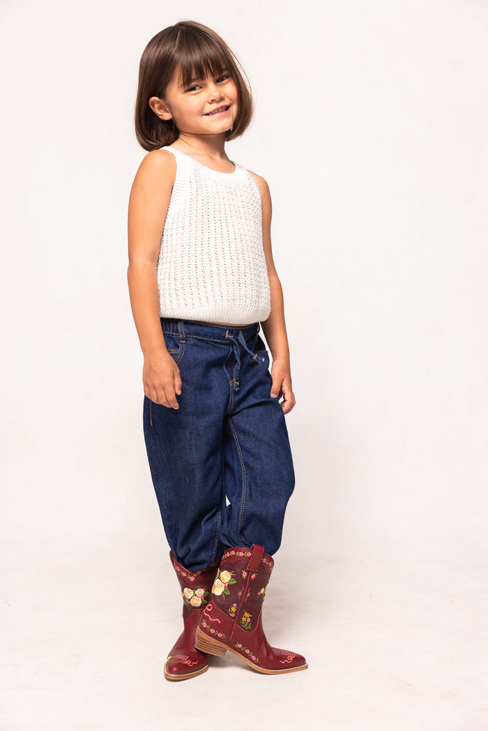 Child wearing a white sleeveless top, blue jeans, and red boots with gold patterns on a white background