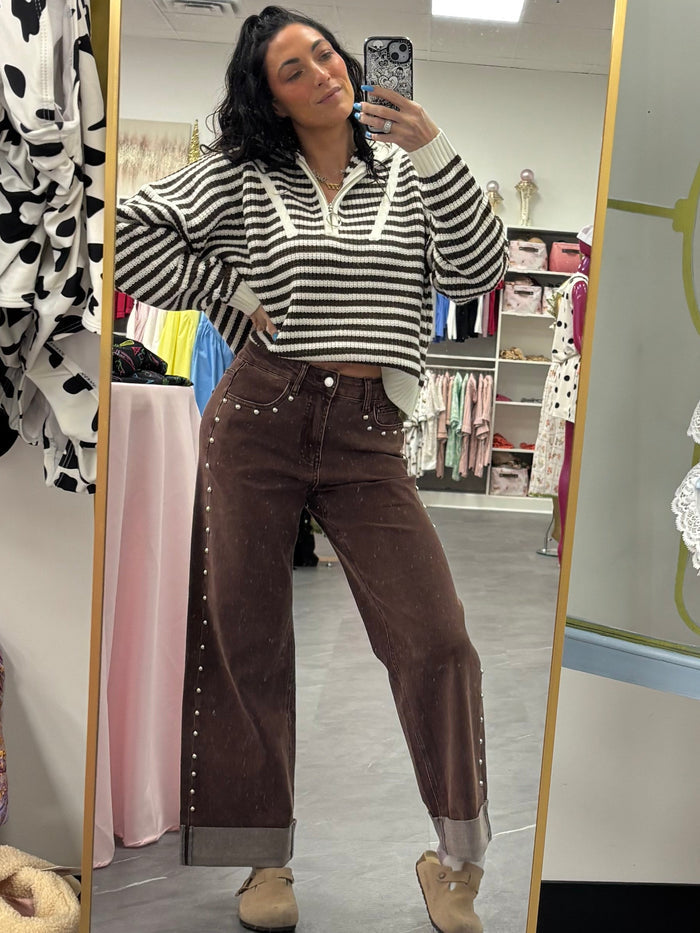 A woman takes a mirror selfie in a boutique showing a striped long-sleeve top and brown pants, while a white lace lingerie bodysuit is displayed on a pink hanger to the right.