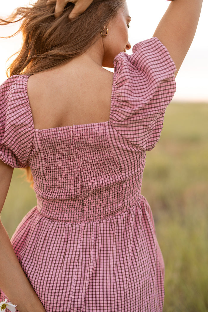 A woman stands in a sunlit field, facing away, wearing the Ivy City Co Monica Dress in Pink & Brown Gingham. Her long hair flows as she lifts one arm, highlighting the dress’s puff sleeves and smocked back.