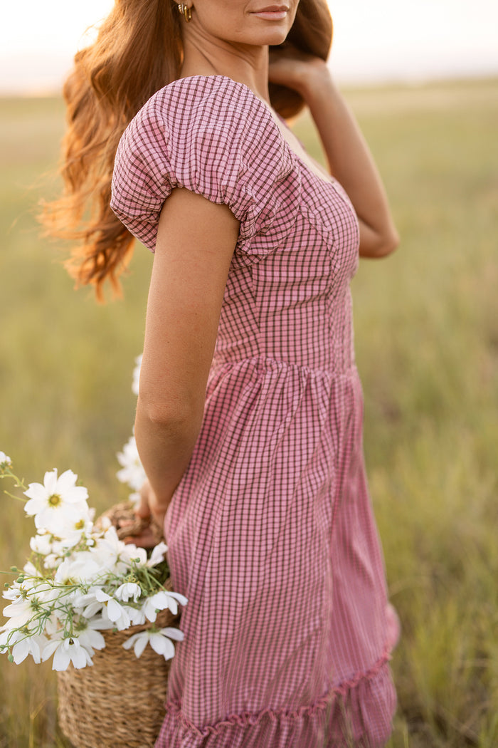 A woman in the Ivy City Co Monica Dress in Pink & Brown Gingham stands in a grassy field, holding a woven basket of white flowers. Her loose wavy hair frames her face as she gazes off to the side, partially out of frame.