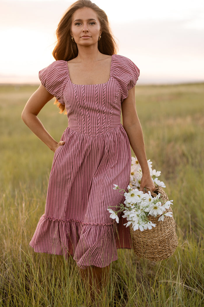 A woman wears the Ivy City Co Monica Dress in Pink & Brown Gingham as she stands in a grassy field at sunset, holding a woven basket filled with white flowers.