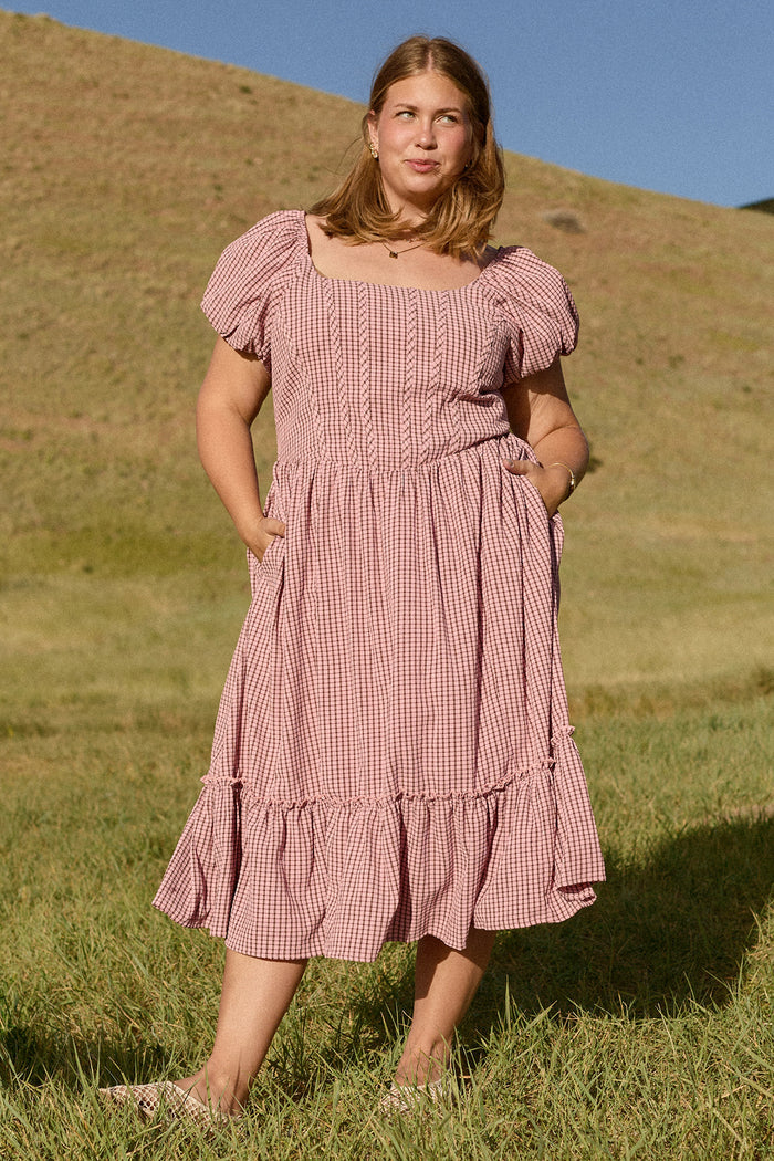 A woman stands barefoot on grass before a hillside, wearing the Monica Dress in Pink & Brown Gingham by Ivy City Co. The midi dress features puff sleeves, a ruffled hem, and pockets. She looks off to the side against a blue sky backdrop.