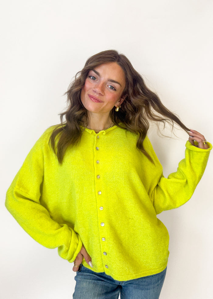 A woman wearing a bright neon yellow-green fuzzy button-up cardigan sweater and blue jeans, posing against a plain white background.