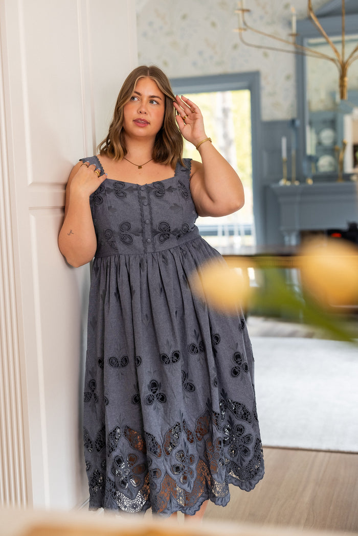 A woman with light brown hair wears the Ivy City Co Penny Tank Midi Dress in Embroidered Chambray. Indoors, she touches her hair and gazes thoughtfully, surrounded by a softly decorated background.