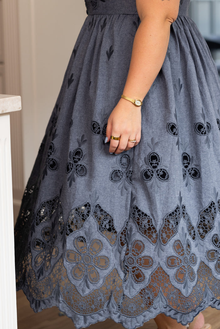 A person wears the Ivy City Co Penny Tank Midi Dress in embroidered chambray, featuring gray cotton fabric, intricate floral lace cutouts near the hem, and eyelet details. They accessorize with a watch and rings on their left arm while standing indoors by a counter.