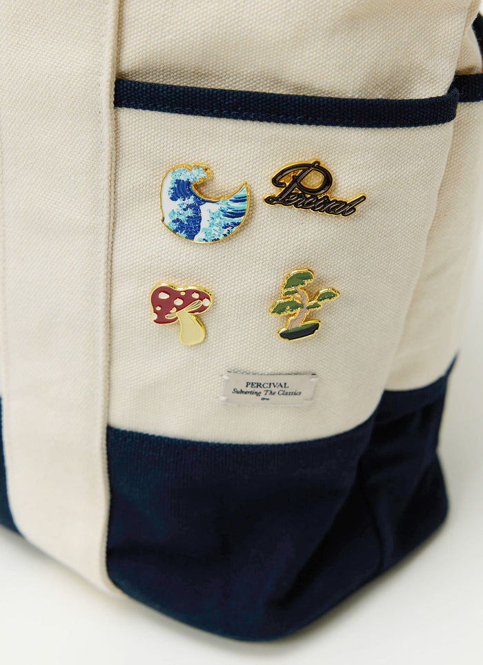 A close-up of the collectable Percival Bonsai, Wave, Mushroom, and script logo pins on a cream canvas tote bag.