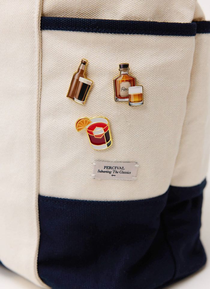 A close-up of the Percival Old Fashioned and other drinks-themed pin badges, pinned onto a cream canvas tote bag.